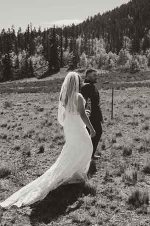 Black and white photo of a bride and groom walking across a mountain meadow.