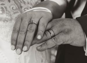 Close-up black and white photo of wedding rings and hands resting on lace fabric.