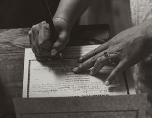 Hands with wedding rings sign an official document in a vintage black and white photograph.