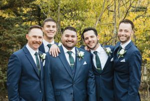 Wedding party wearing matching navy blue suits and boutonnières pose together outdoors with autumn foliage in background.