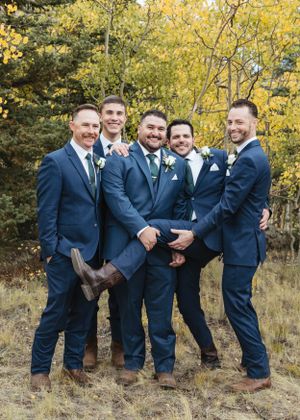 A wedding party in navy blue suits poses together on a hillside with golden autumn foliage in the background.