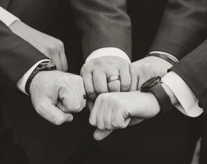 Three hands wearing formal suits and wedding rings join together in a black and white photograph.