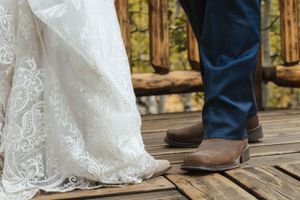 Close-up of a wedding dress hem with delicate lace detail next to brown cowboy boots on wooden deck.