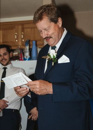 Wedding guest in navy suit with white boutonniere reads from a paper during indoor reception.