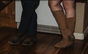 A pair of brown suede boots and black shoes standing on hardwood flooring next to a white baseboard heater.