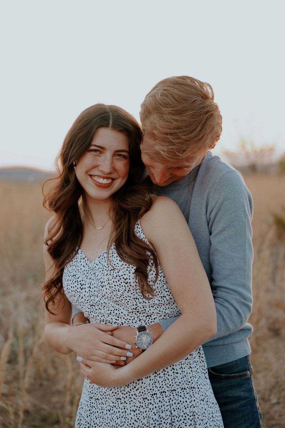 A smiling brunette woman in a floral dress embraced by a blonde-haired man in a golden field at sunset.