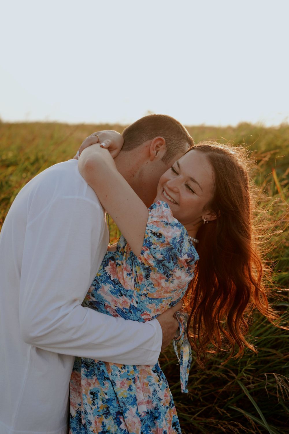 A couple embraces in a golden field at sunset, woman in floral dress smiling joyfully as man kisses her cheek.