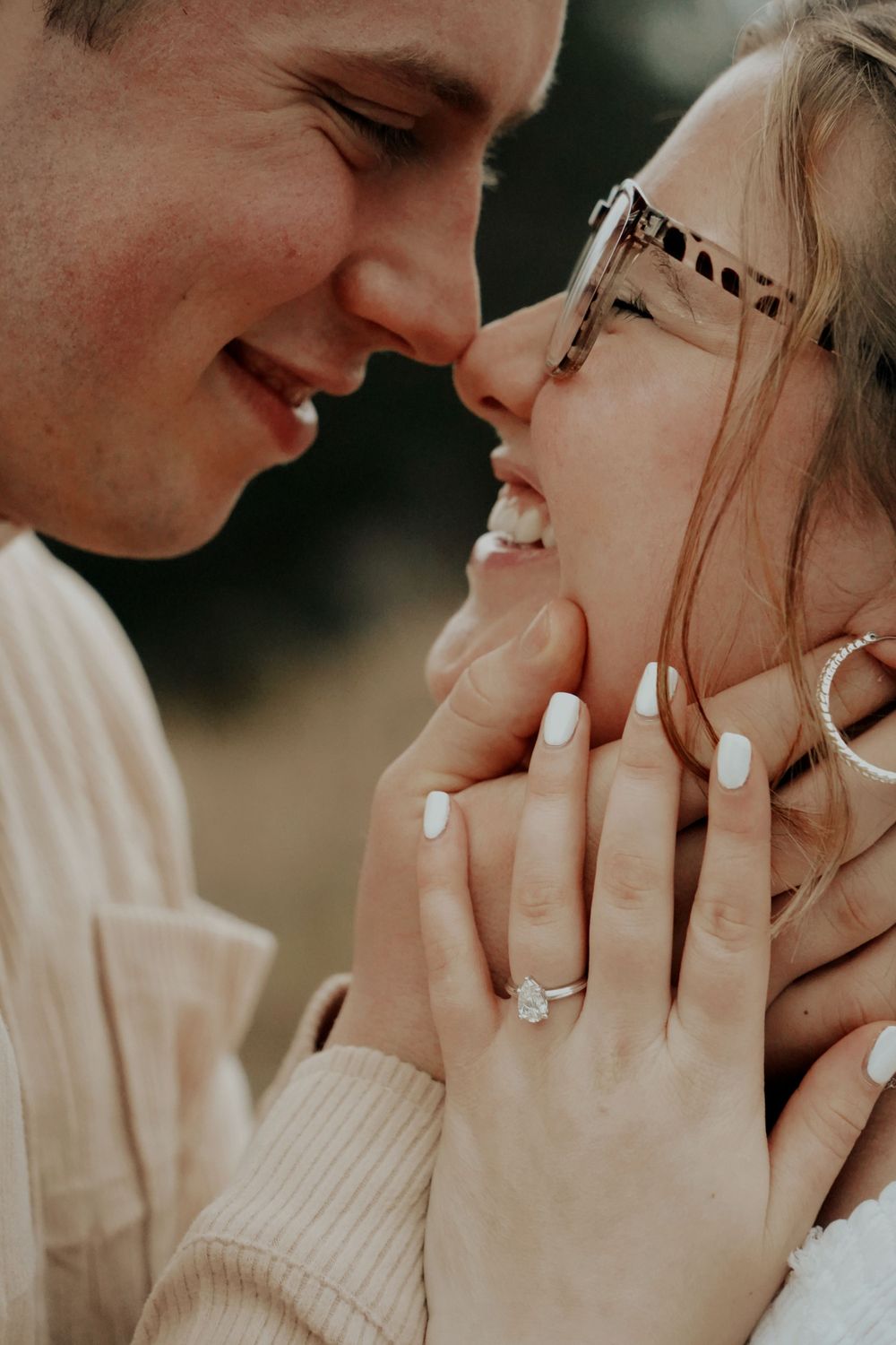 Couple sharing a romantic moment, woman displaying diamond engagement ring with white manicured nails, smiling closely.
