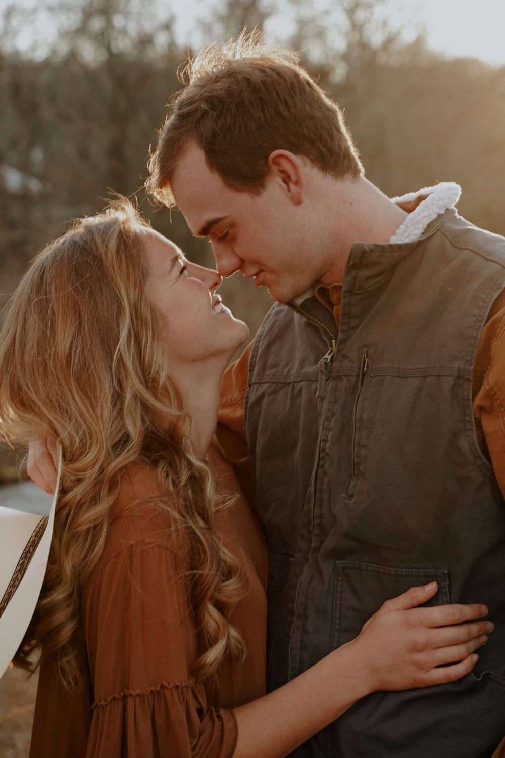 Couple sharing a romantic moment at golden hour, woman in rust dress embracing man in brown vest outdoors.
