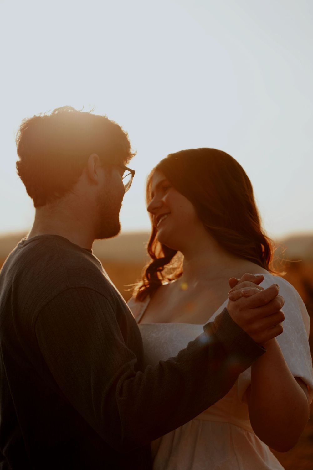 Couple dancing romantically at golden hour sunset, man in glasses holding woman in white dress close.