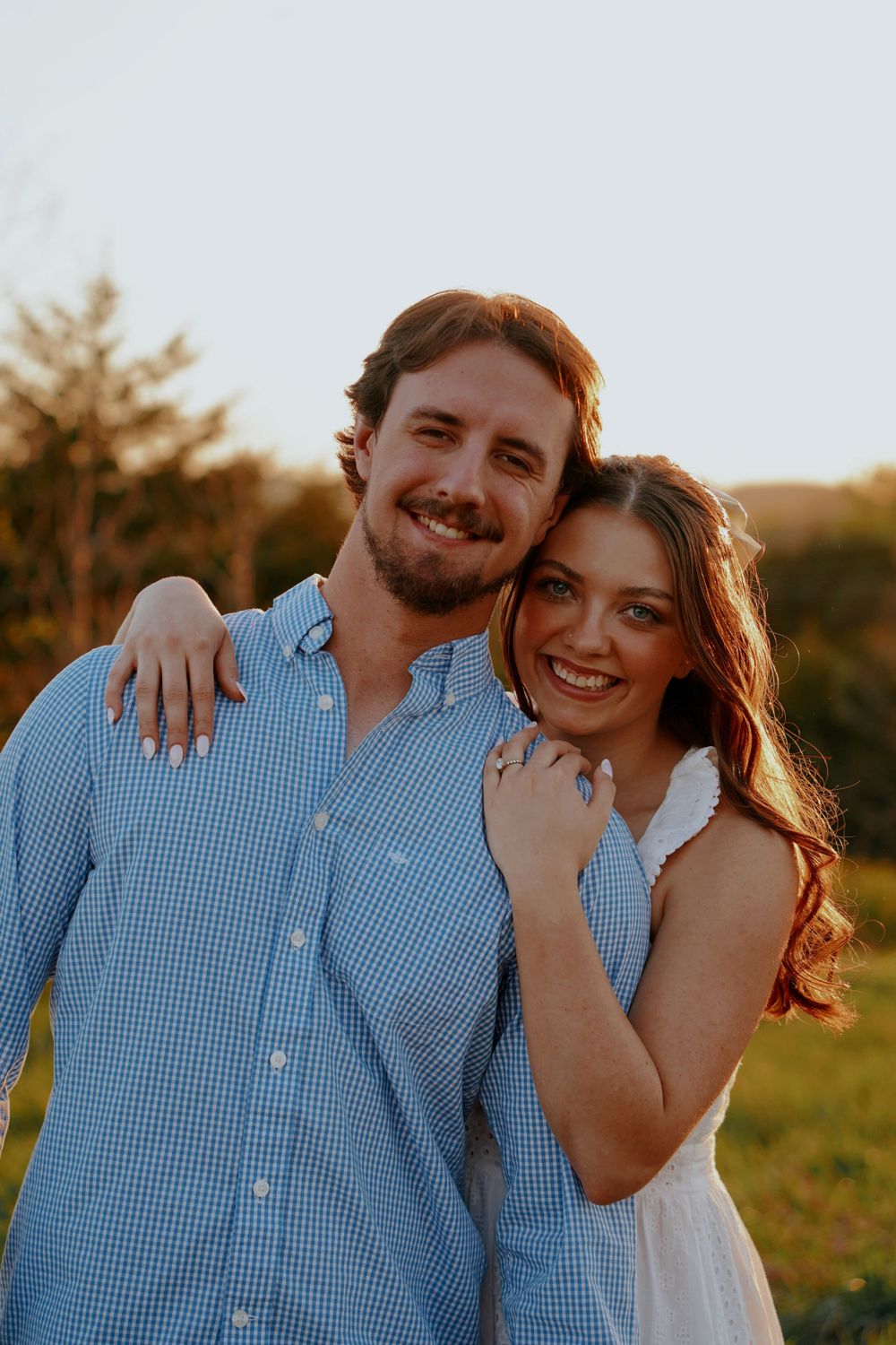 Happy engaged couple smiling at sunset, woman in white dress hugging man in blue checkered shirt.