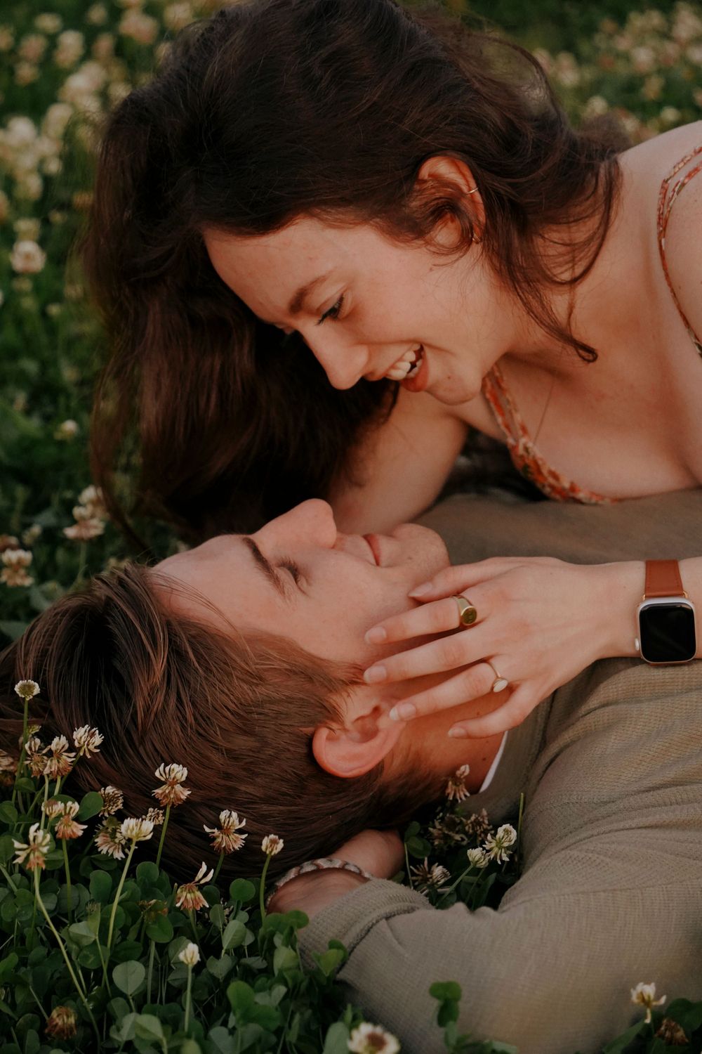 Couple laughing together in a clover field, woman leaning over man lying in wildflowers.