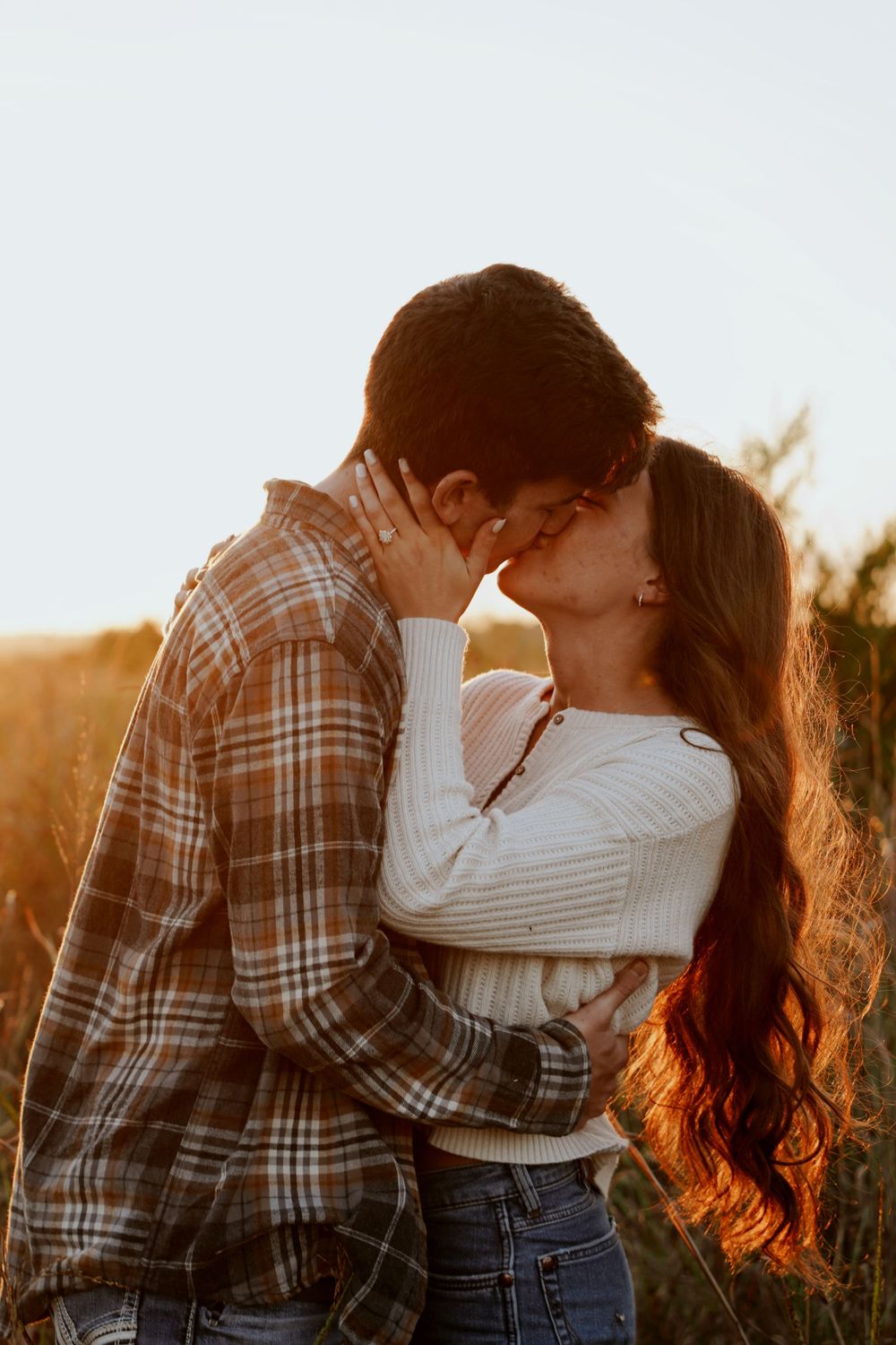 Couple sharing a kiss at golden hour sunset in an open field, woman in white sweater.