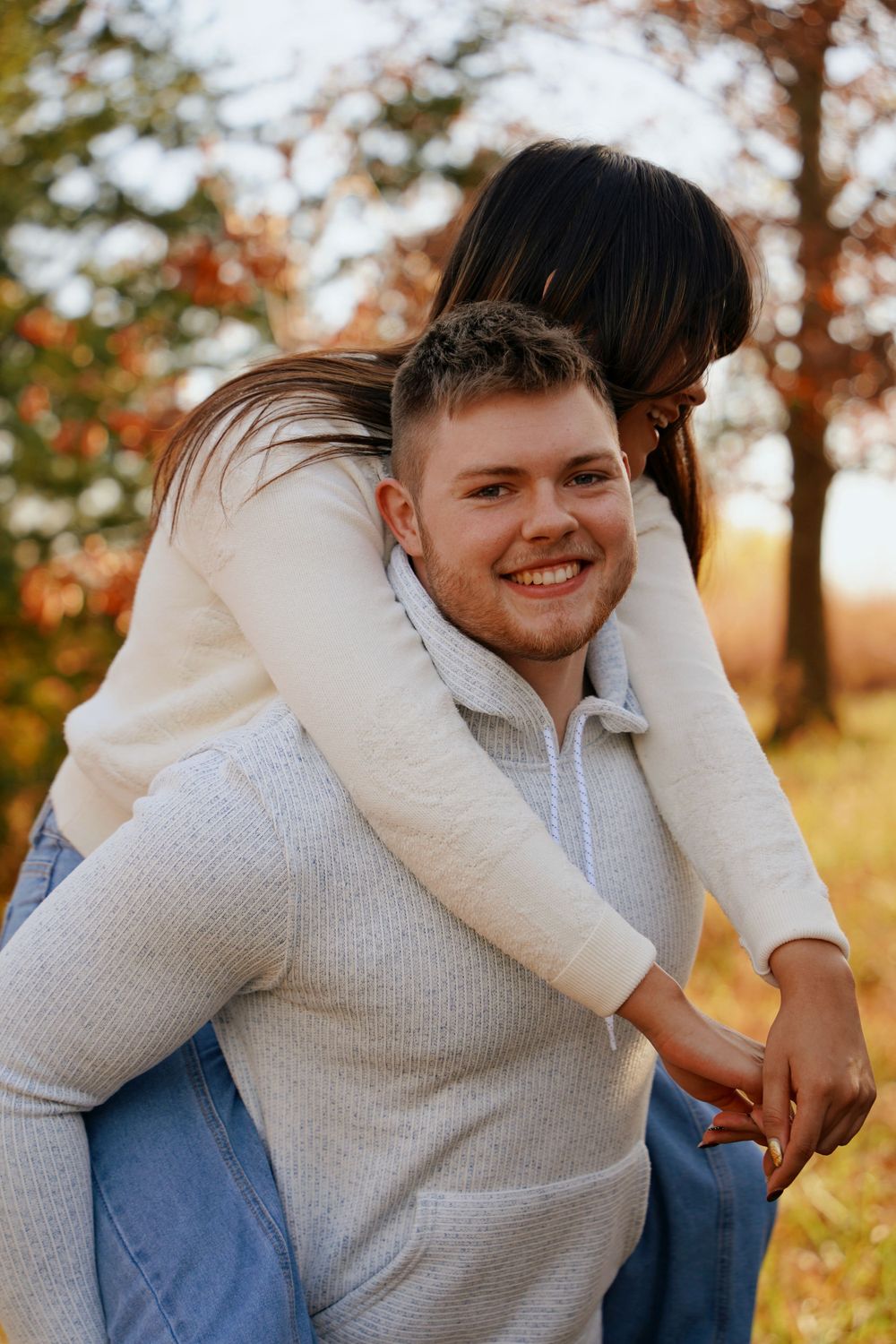 Man giving woman a piggyback ride in autumn park, both smiling and wearing white sweaters.