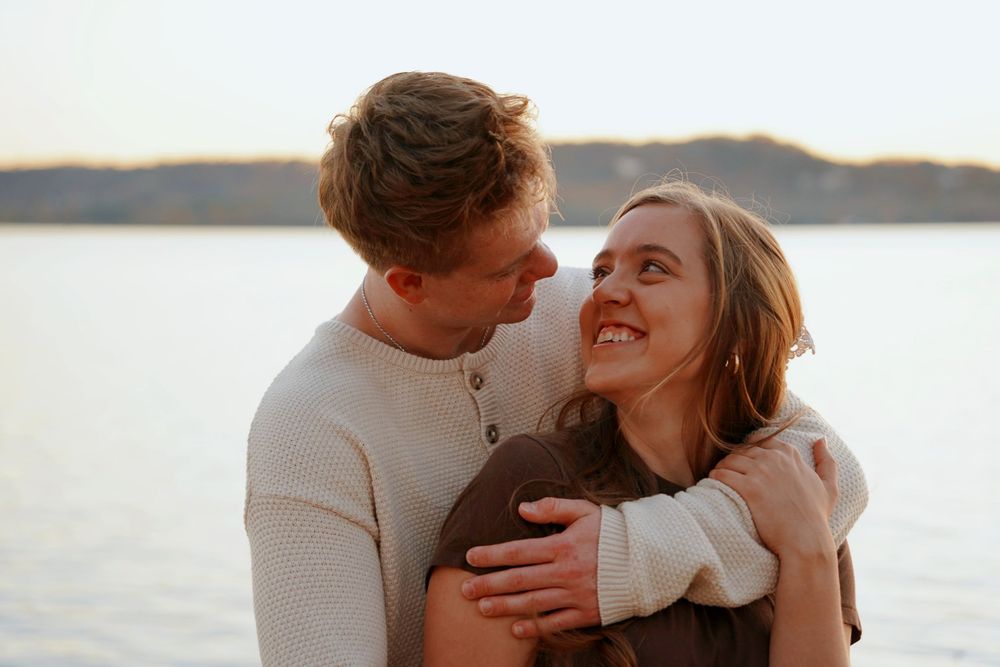 Couple embracing and smiling at each other near a lake at sunset.