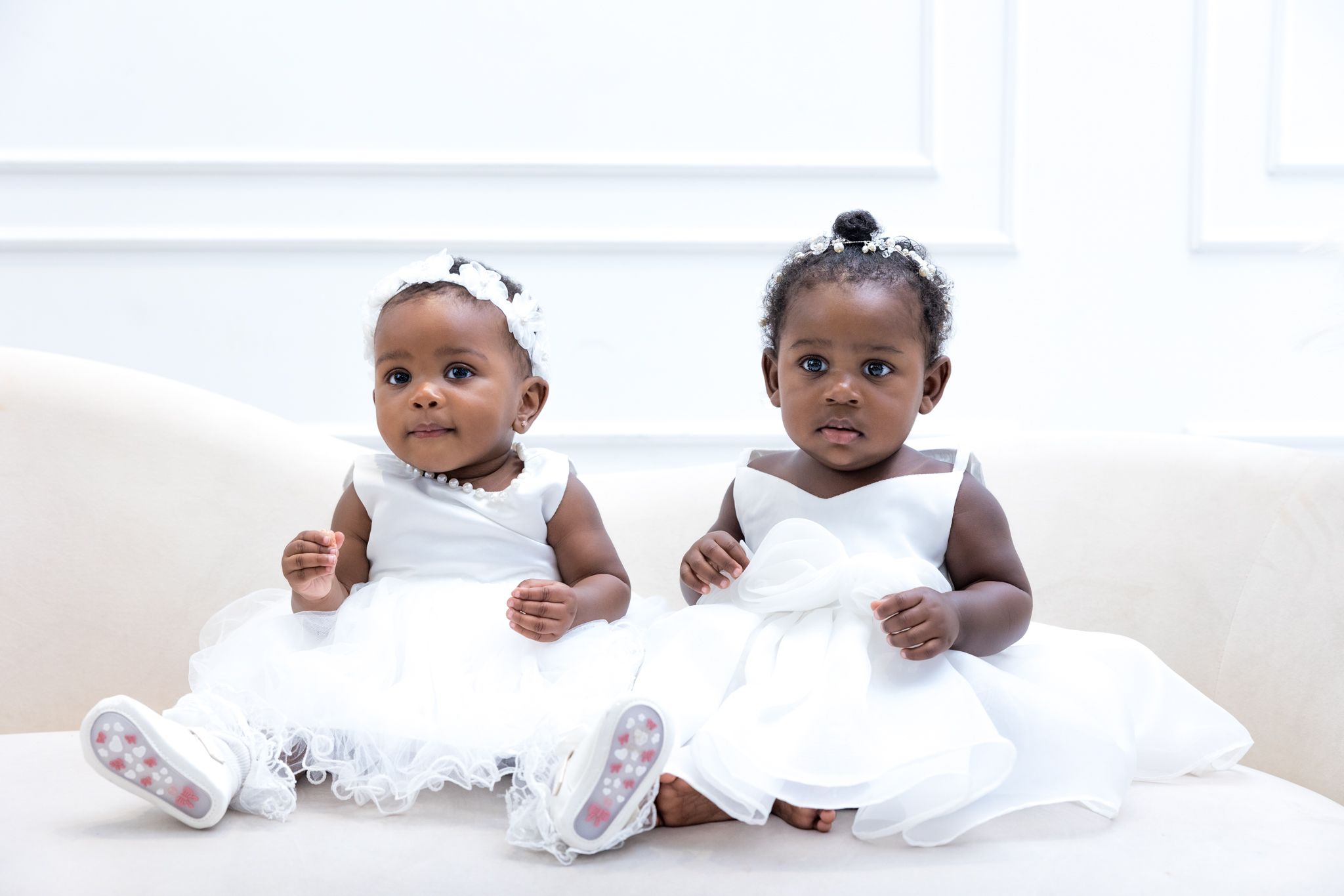 Two babies wearing matching white dresses and bows sit side by side posing for a family photoshoot.