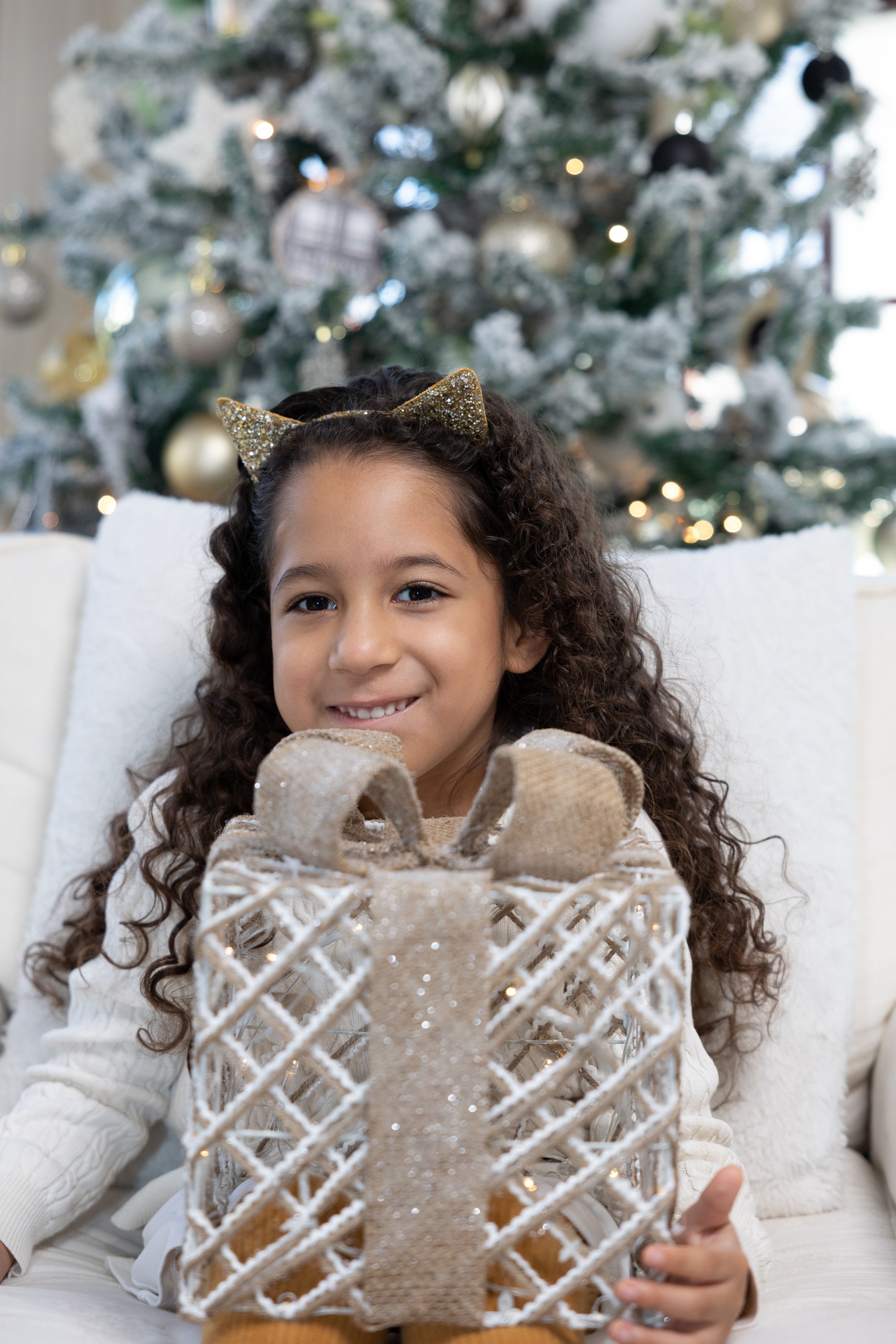 Child in white sweater holds gift while seated by decorated Christmas tree.
