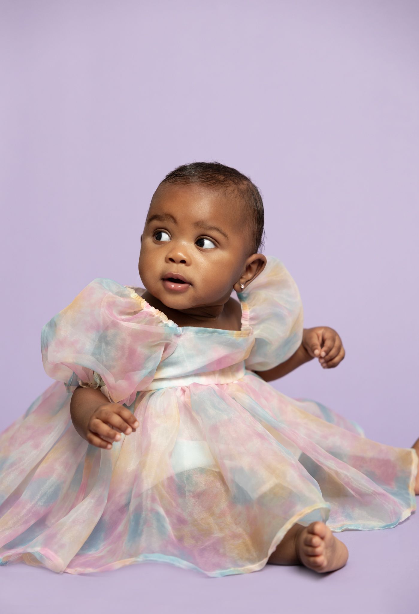 Baby wearing a shimmering pastel dress with ruffled details gazes to the side against a purple backdrop.