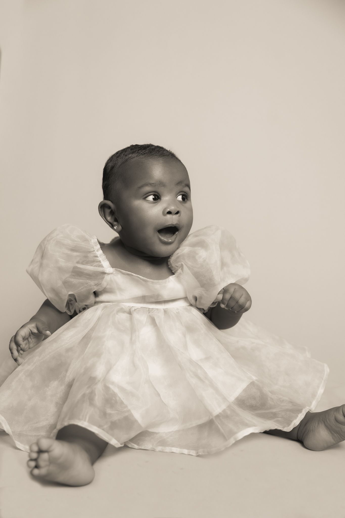 A baby in a white frilly dress sits on the floor with an excited expression in this sepia-toned portrait.