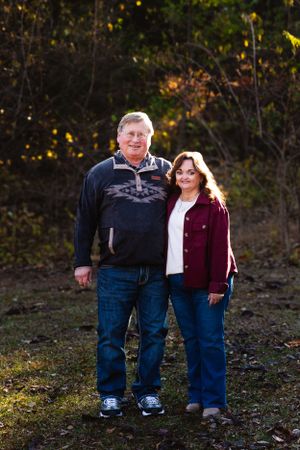 Couple stands together smiling in casual attire against fall foliage backdrop.
