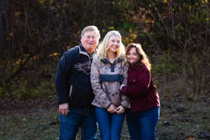 Three people pose together outdoors wearing casual clothing in front of autumn trees.
