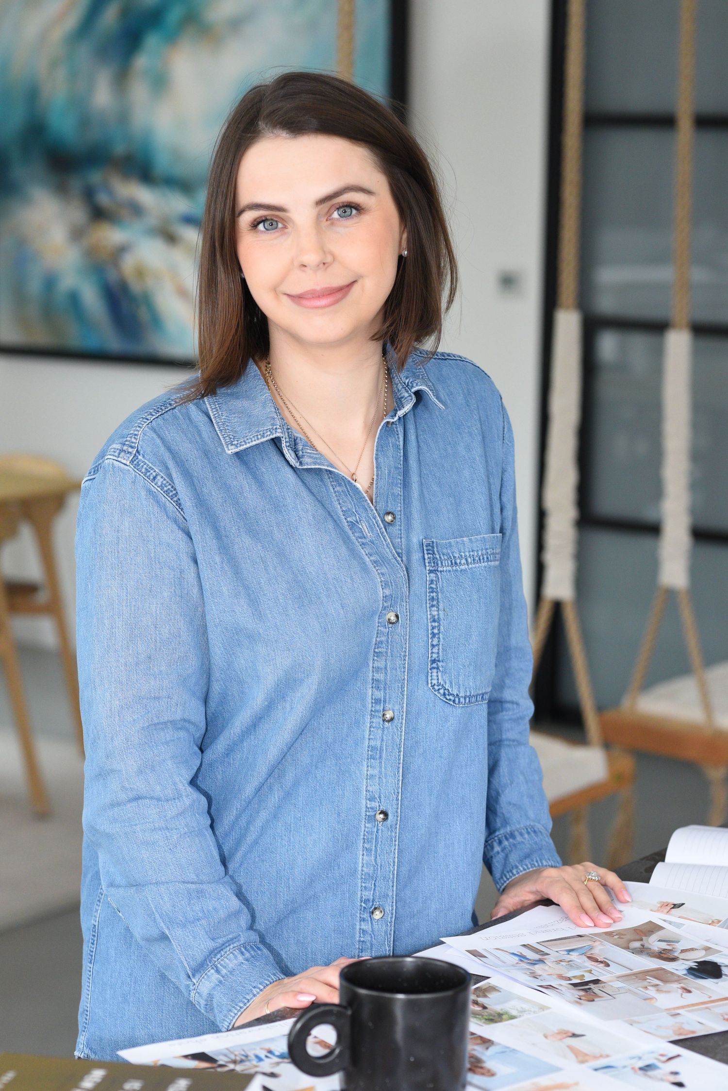 Professional wearing denim button-up shirt stands at desk with coffee mug and paperwork in bright modern office space.