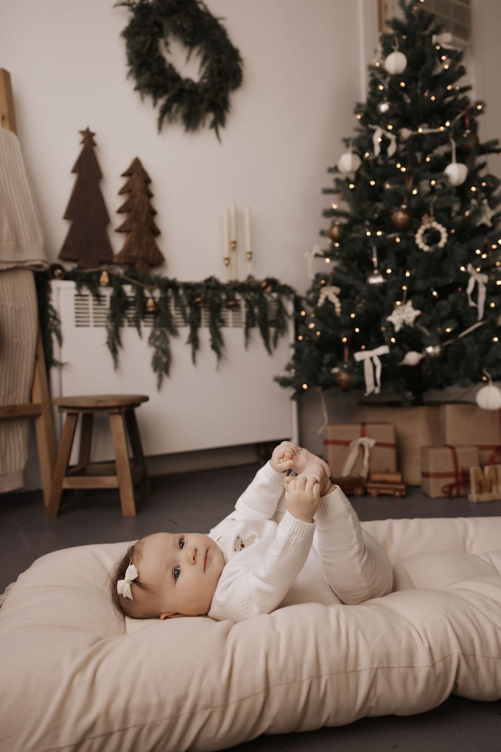 Baby in white outfit lying on cushions with Christmas decorations and tree in background.