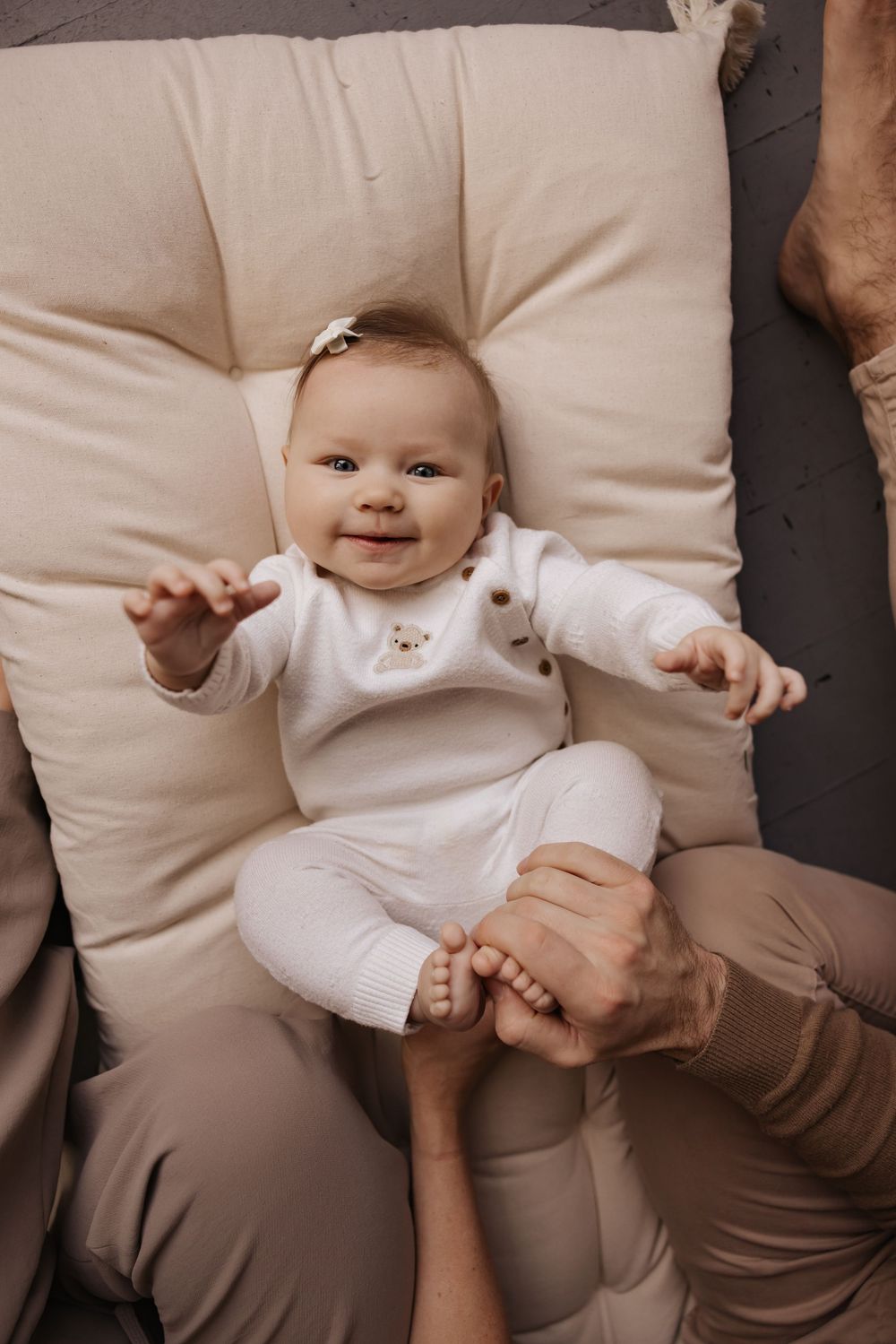 Baby in white outfit playing and smiling on cream cushions.