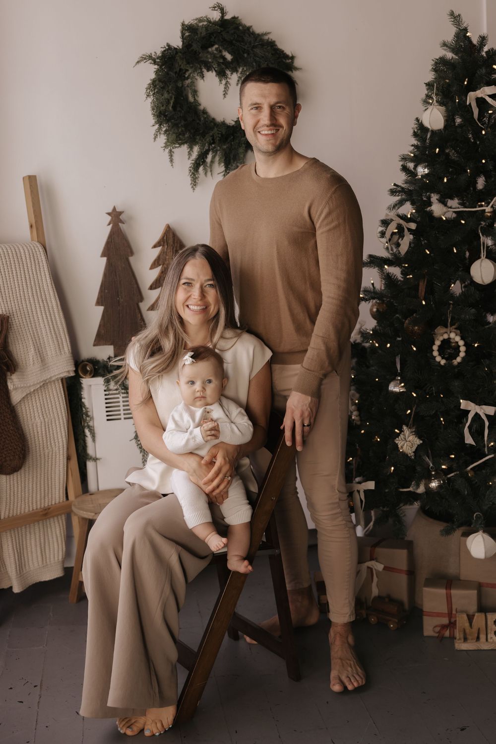 Family in neutral tones posing together by Christmas tree and decorative wooden trees.