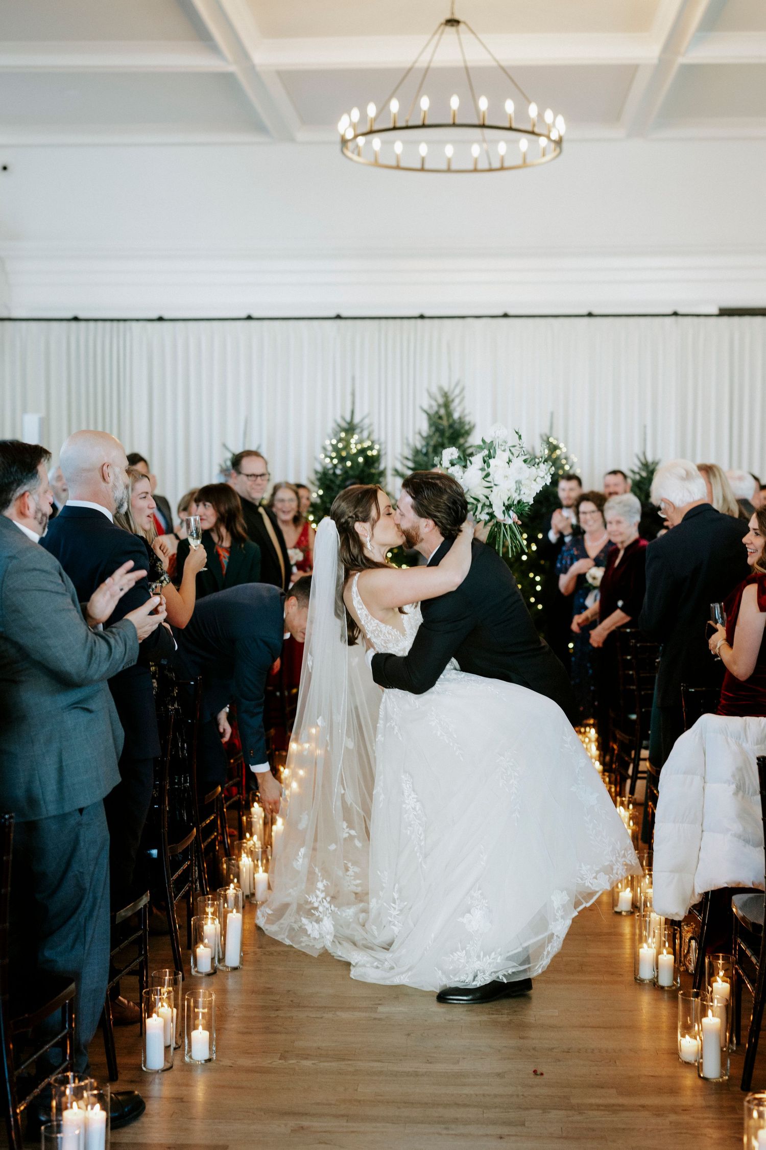 Romantic wedding couple performs a dramatic dip during their first dance in an elegant white ballroom with glowing candles.