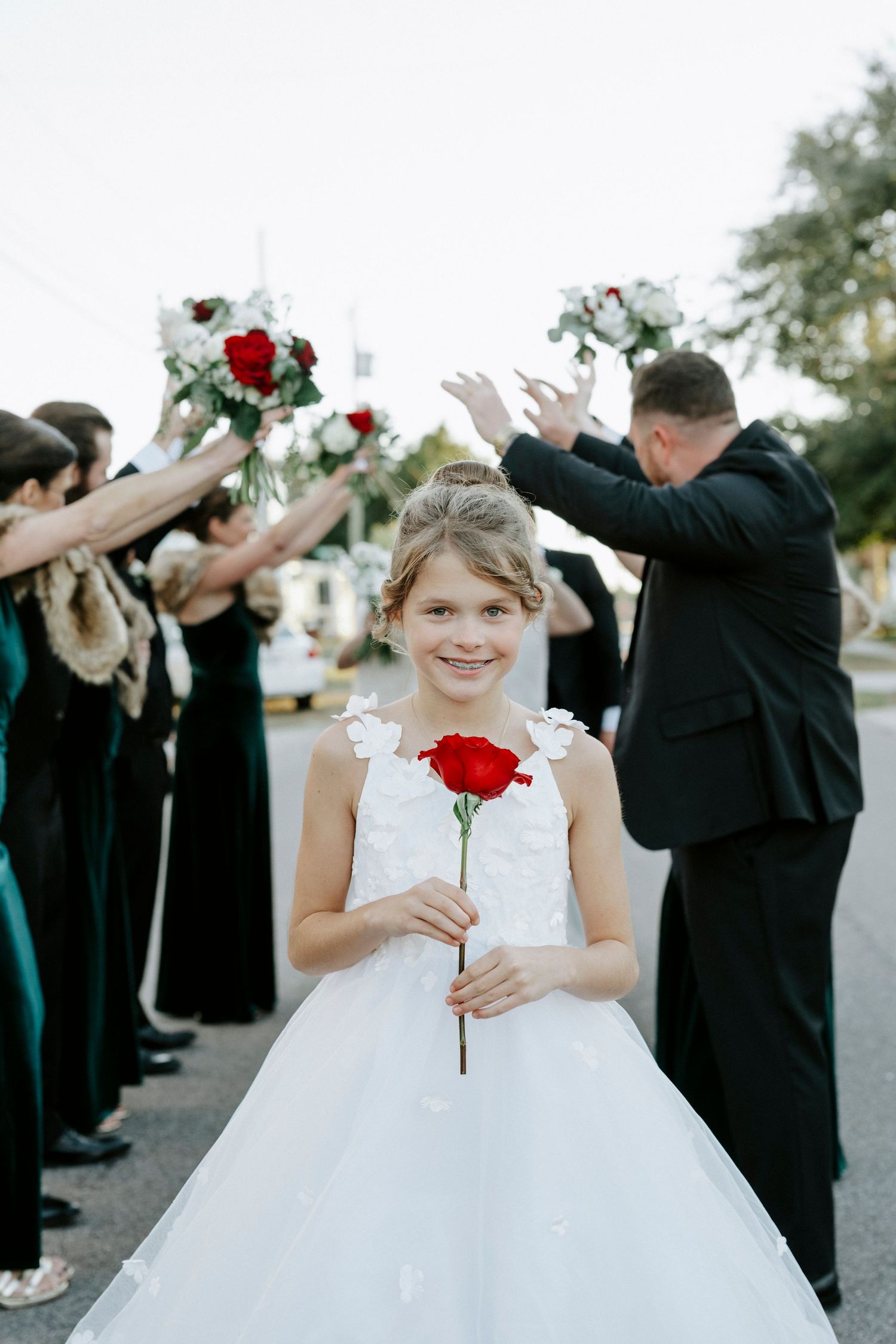 Wedding party forms a tunnel with raised arms while flower girl in white dress holds red rose at outdoor celebration.