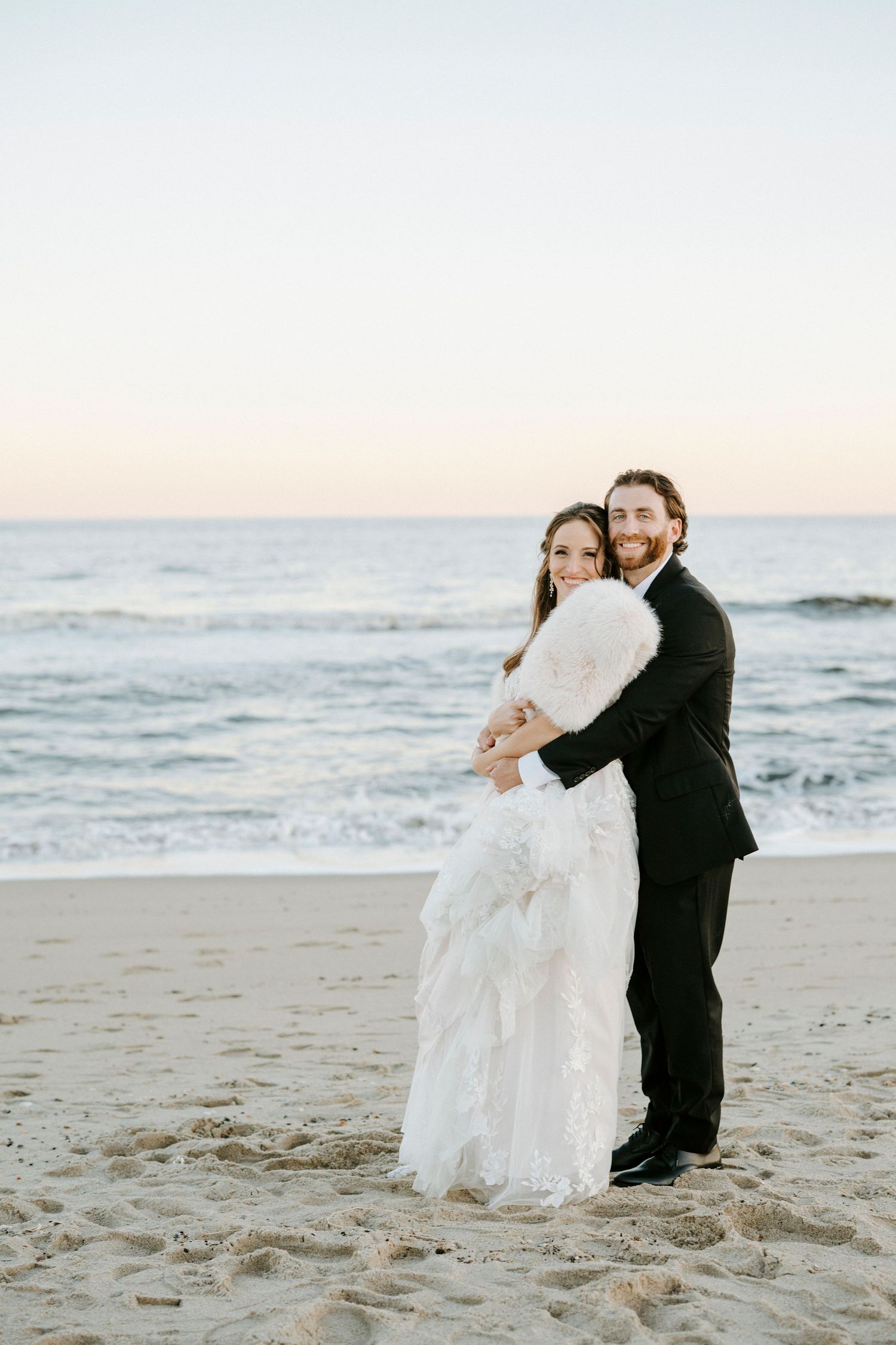 A couple in wedding attire sits together on a sandy beach at sunset with ocean waves in the background.