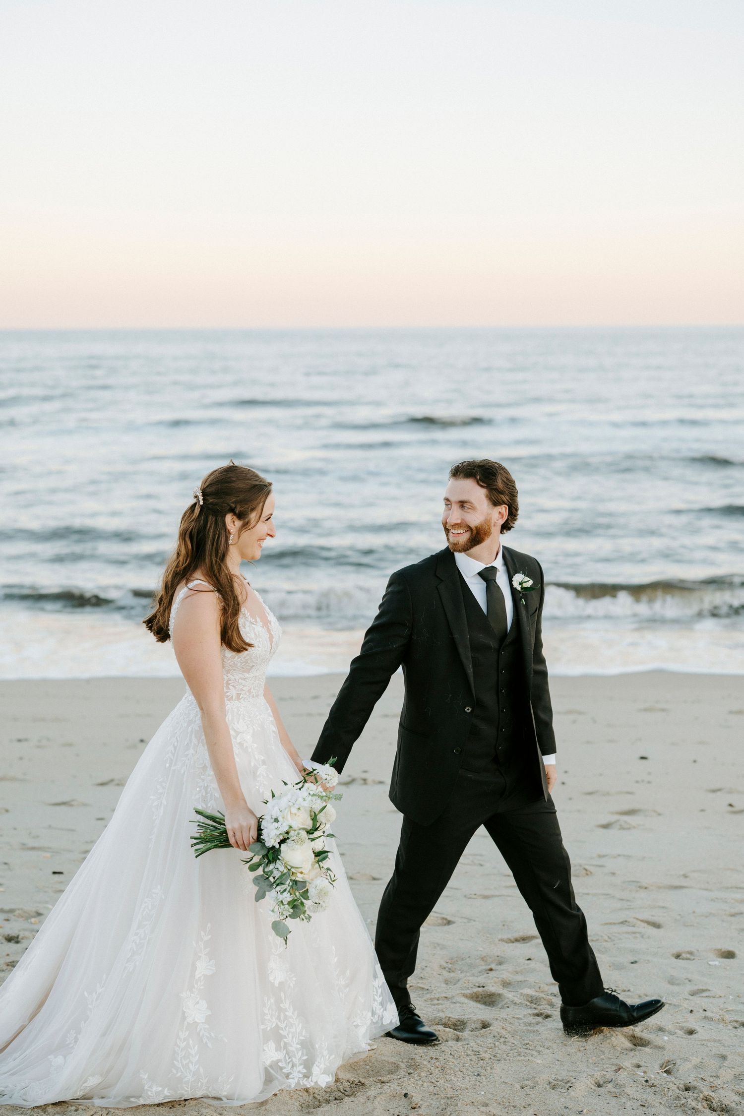 Newlywed couple shares playful moments on a beach at sunset while dressed in wedding attire.