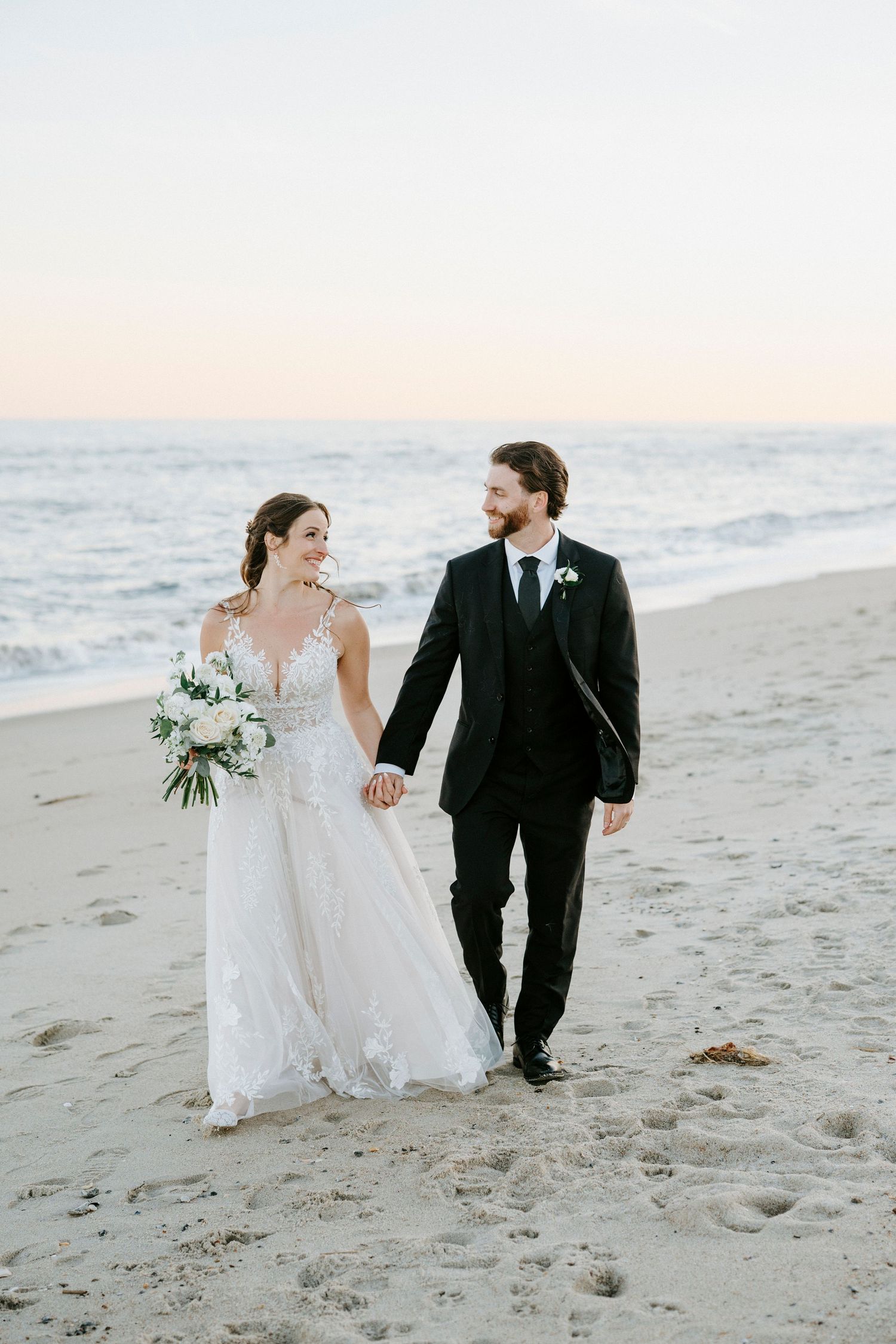 A couple walks together on a sandy beach at sunset wearing wedding attire.