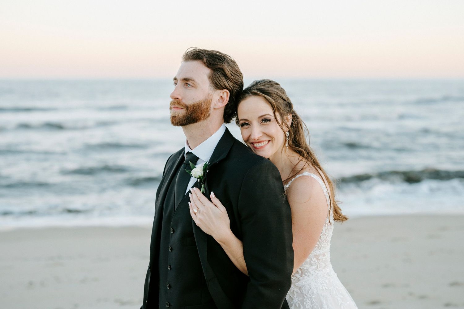 A couple in wedding attire embraces on a beach at sunset with ocean waves in the background.