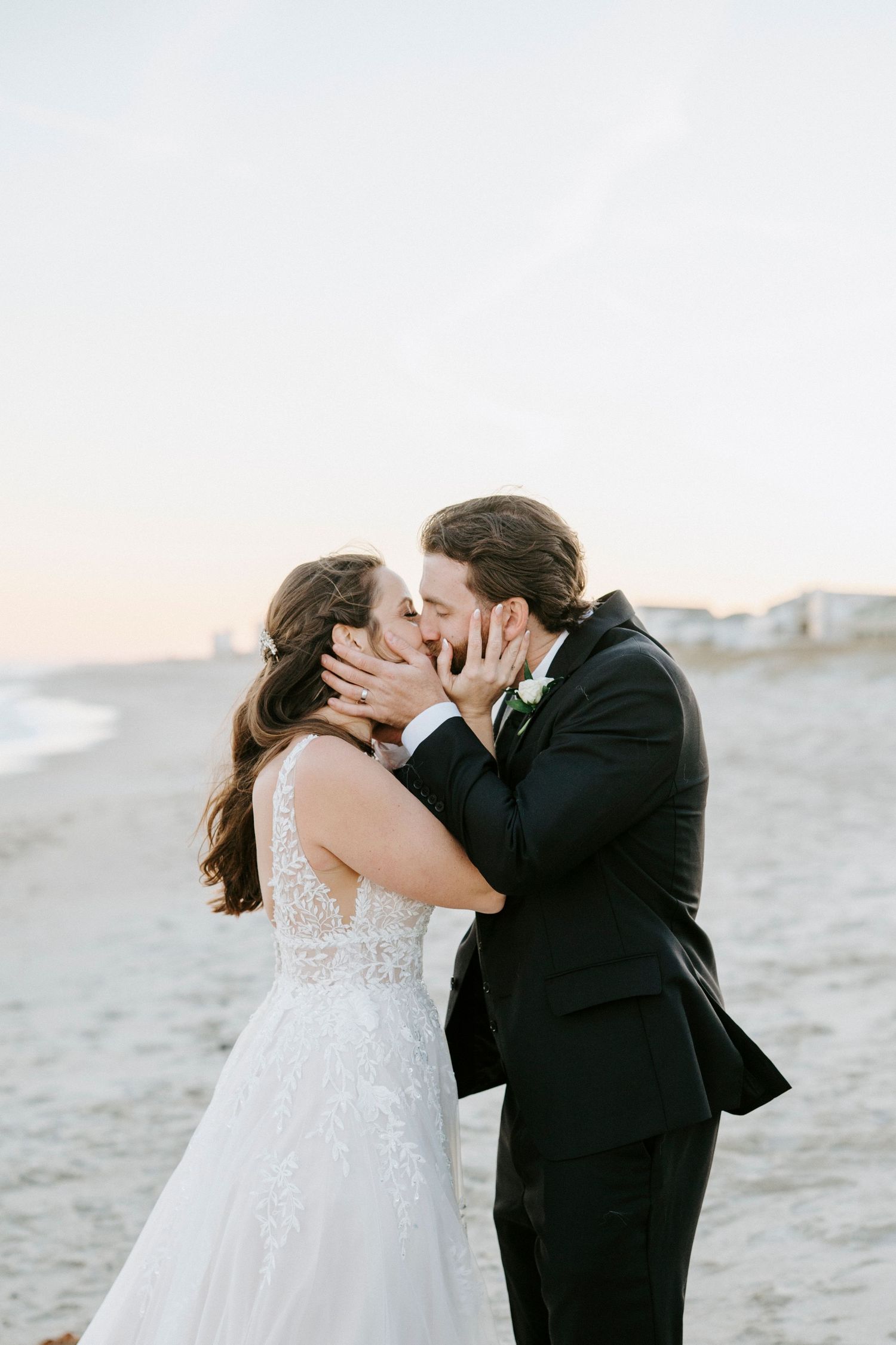 A couple shares a romantic kiss on a sandy beach during their wedding photoshoot at sunset.