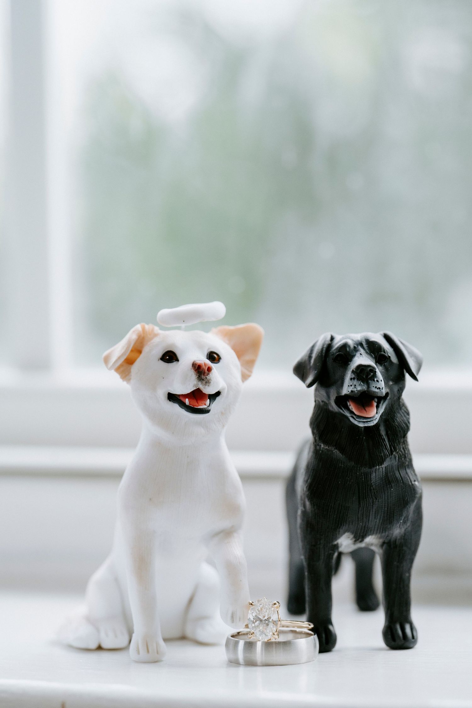Two dog figurines, one black and one white, displayed with wedding rings on a windowsill.