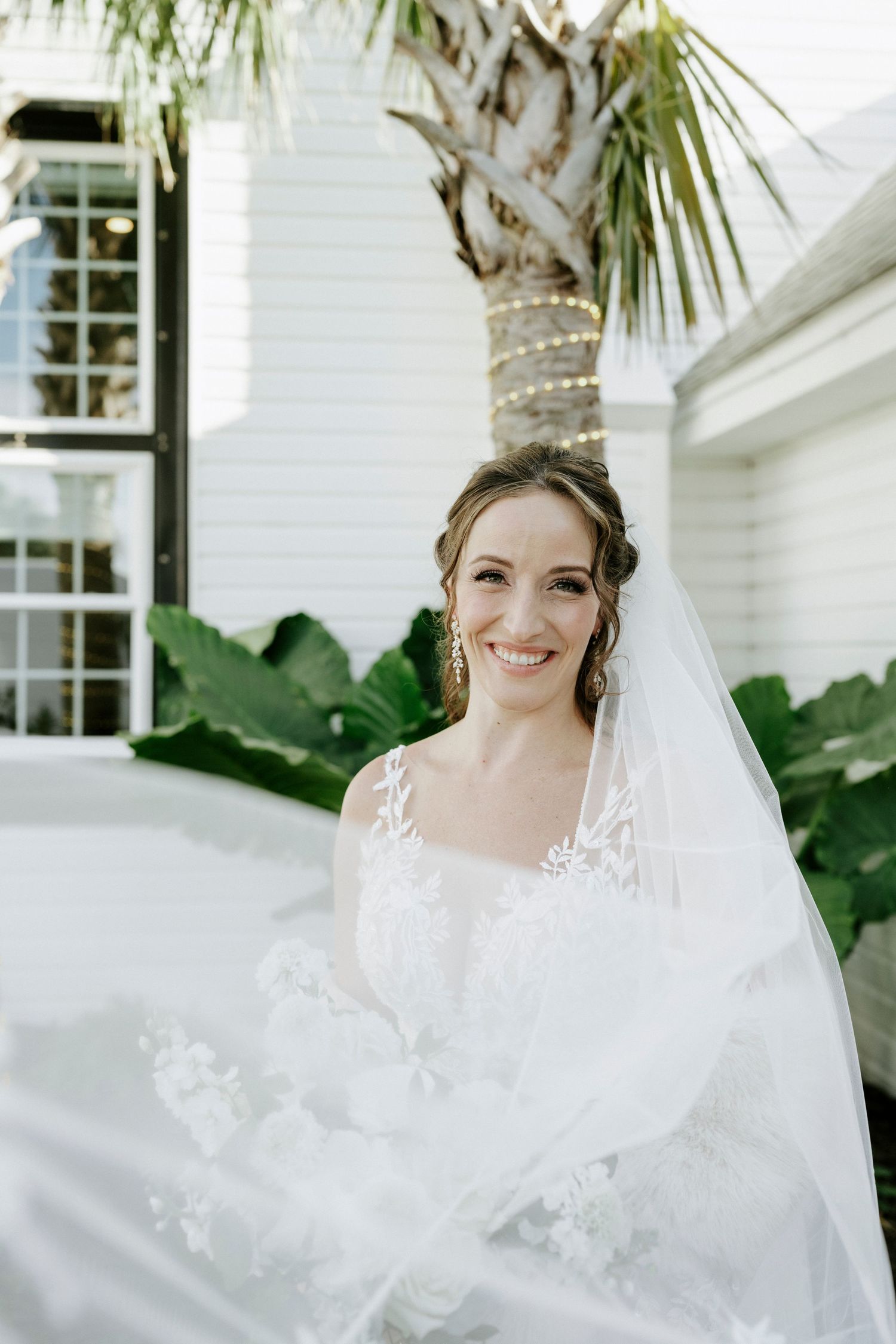 A beautiful white wedding dress with delicate lace details flows elegantly against a backdrop of tropical palm leaves.