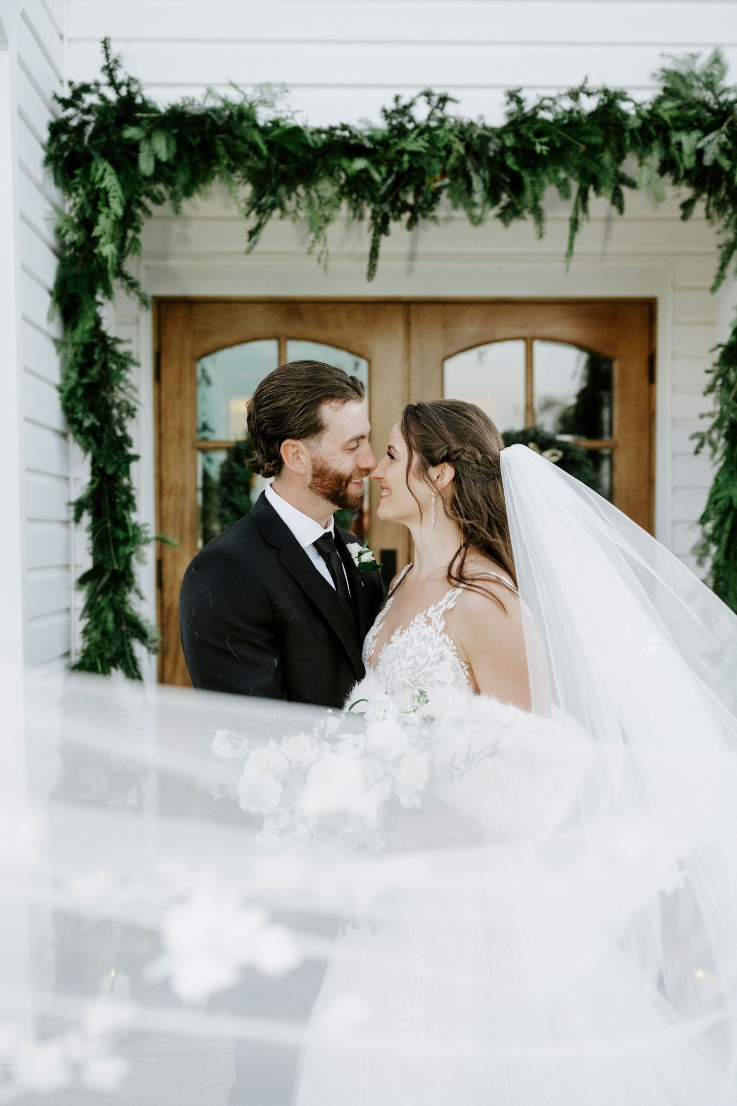 A romantic wedding couple shares a tender moment against a white building with greenery and windows.