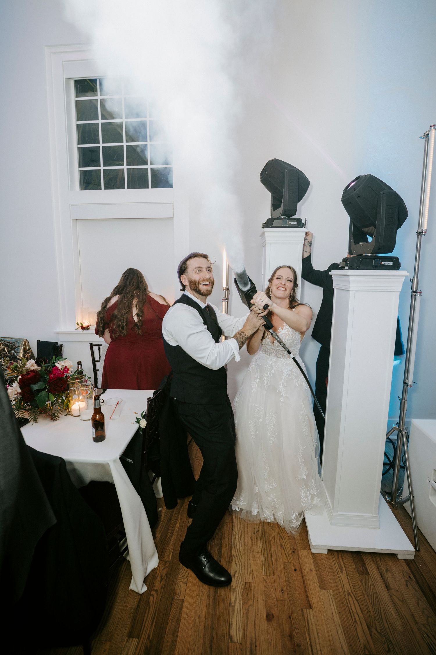 Wedding party members dance and celebrate in a bright indoor venue with wooden floors and white walls.