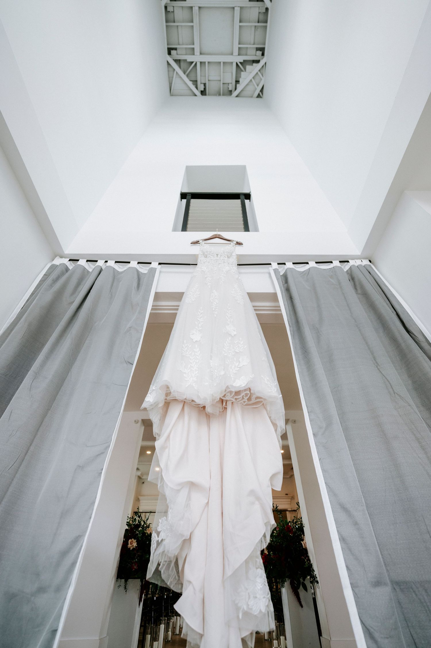 A white wedding dress hangs from a ceiling fixture in a bright room with gray walls and hardwood floors.