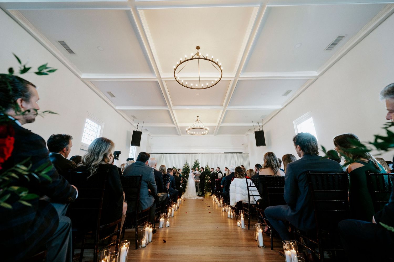 Wedding ceremony in a bright white venue with guests seated in rows beneath elegant chandeliers and potted plants.