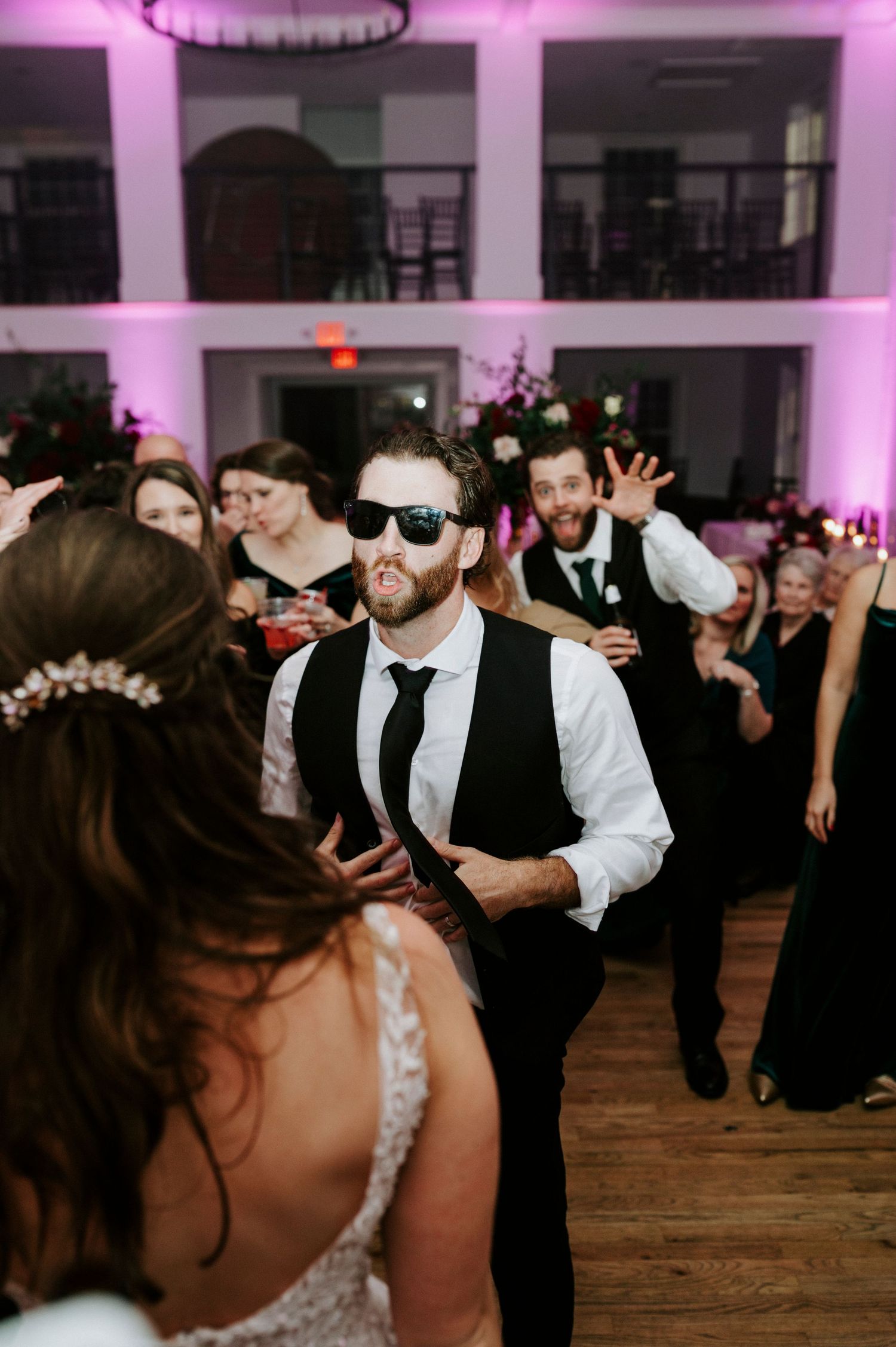 Wedding reception guests dancing and celebrating on a dance floor illuminated by pink lighting.