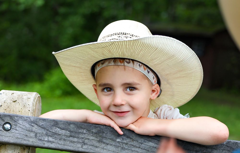 A young child wearing a wide-brimmed straw hat leans on a wooden fence in a sunny outdoor setting.