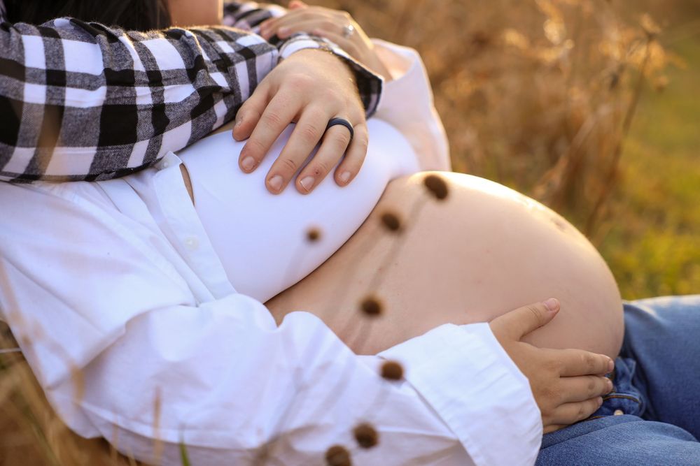 A pregnant woman in a white dress and plaid shirt cradles her baby bump in a golden sunlit outdoor setting.