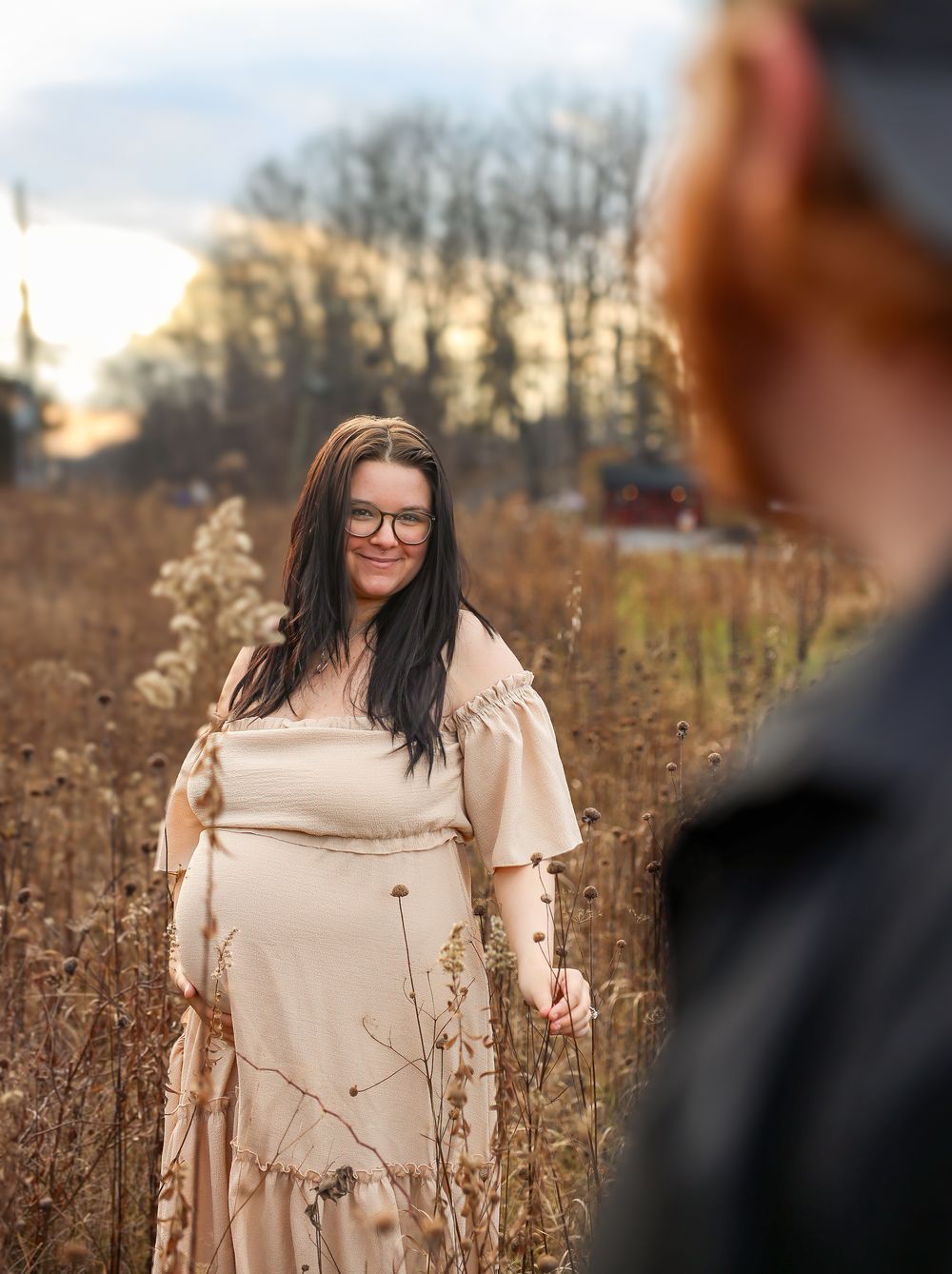 A woman in a beige dress stands in a field of dried autumn grasses at sunset with trees silhouetted in the background.