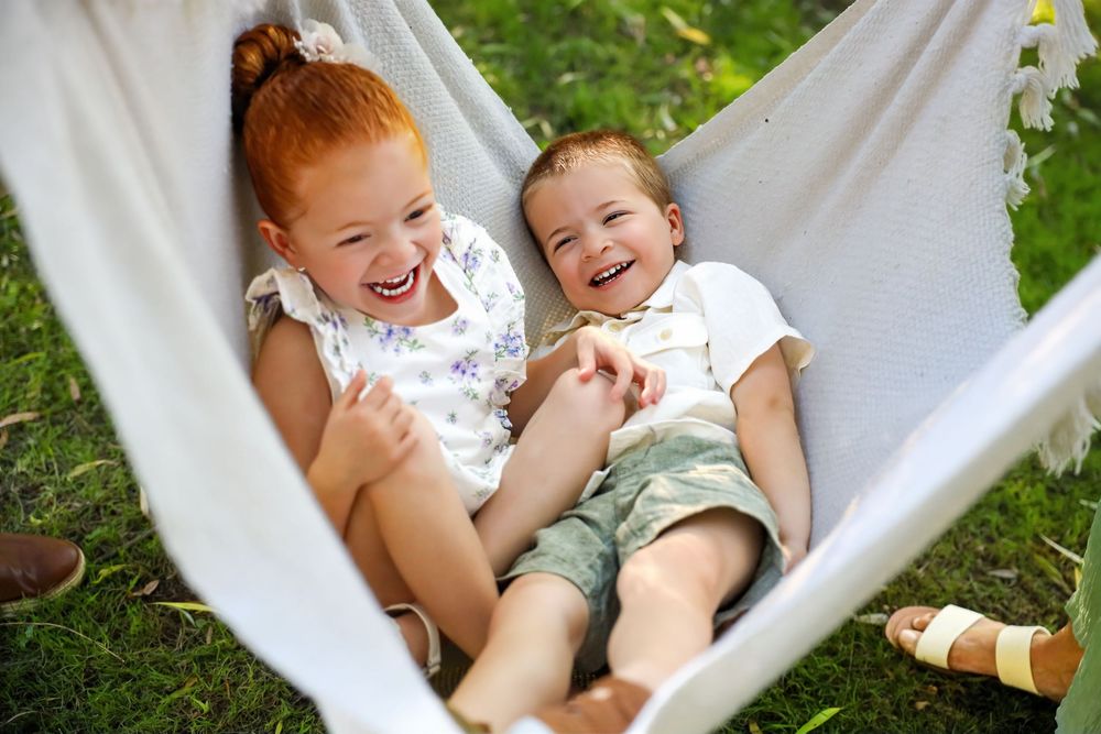 Two children share joyful laughter while relaxing together in a white hammock outdoors on a sunny day.