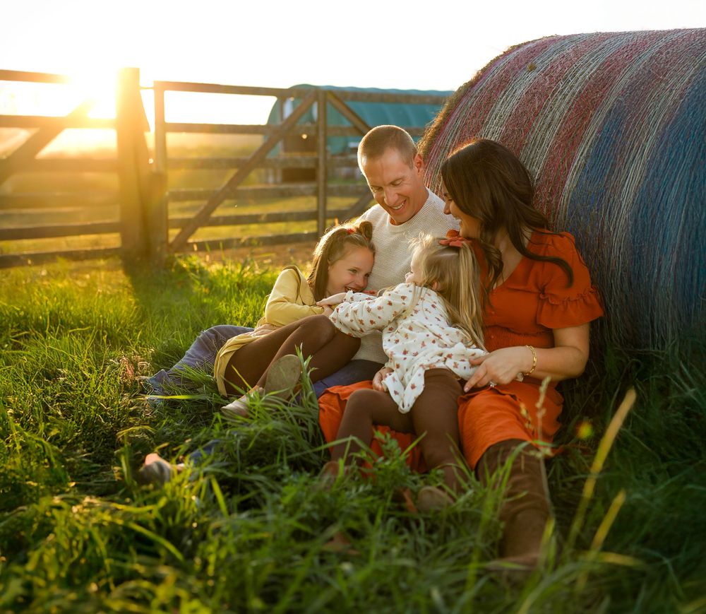 Couple sitting together in tall grass at sunset with hay bales creating a romantic golden hour atmosphere.