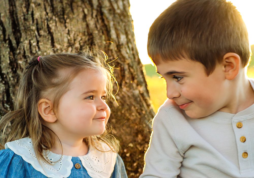 Two children share a sweet moment together while sitting by a large tree trunk in warm sunlight.