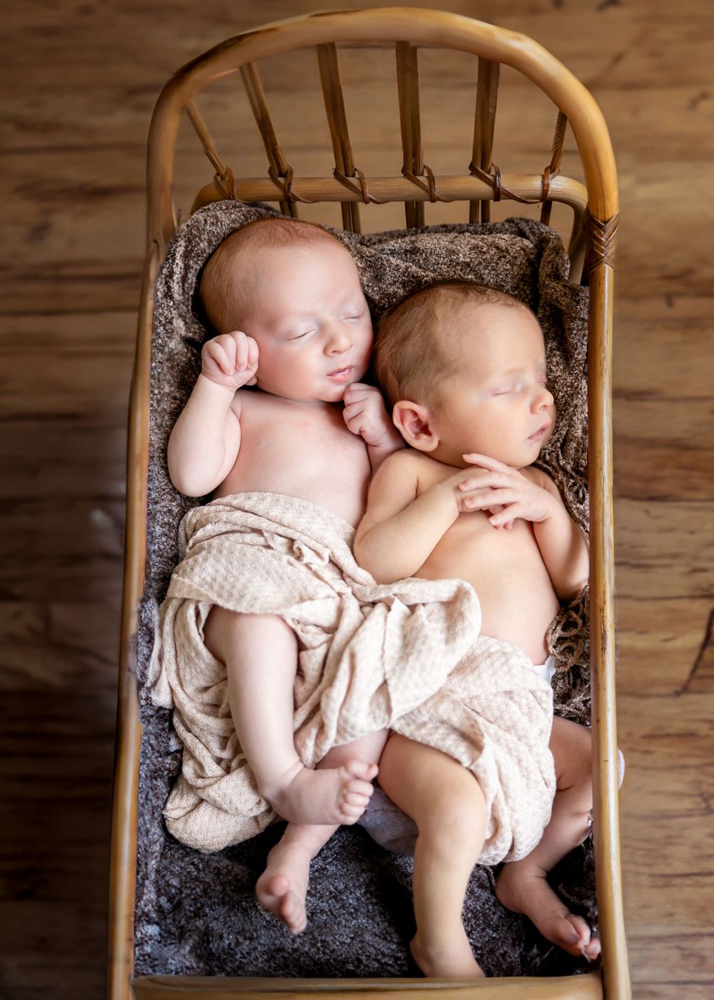 Two sleeping babies wearing white outfits snuggle together on a dark cushion in a wooden chair.